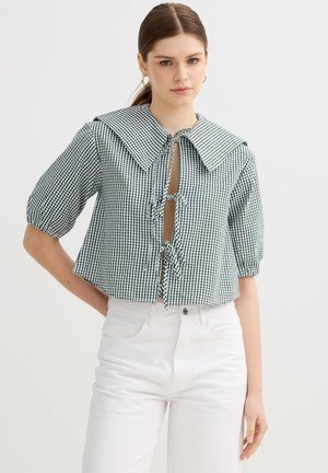 Joven mujer con blusa de manga corta a cuadros verdes y blancos con cuello grande y cierres con lazos, combinada con pantalones blancos de talle alto.