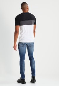 Camiseta de bloques de color negro, gris y blanco; manga corta; material de algodón suave. Se lleva con jeans ajustados azules y zapatos negros.