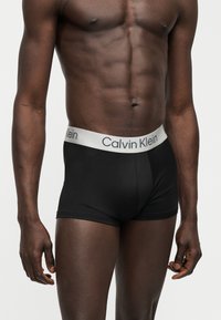 Μυώδης ανδρικός κορμός που φοράει μαύρα εσώρουχα Calvin Klein με λευκή μέση, στέκεται σε απλό φωτεινό φόντο.