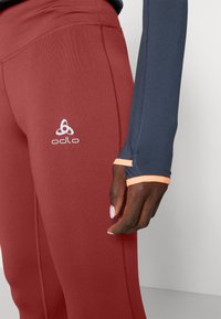 Strak passende sportleggings in een dieprode kleur met slanke stiksels en een Odlo-logo; gecombineerd met een langermige top in marineblauw met oranje accenten.