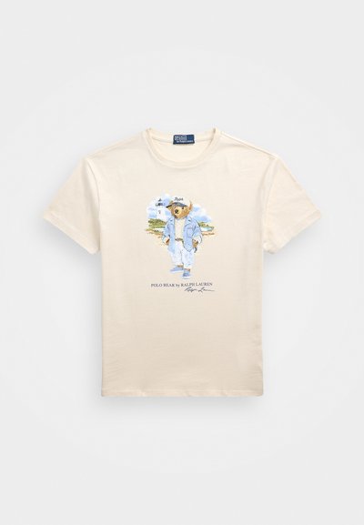 Κρέμα βαμβακερό μπλουζάκι με πολύχρωμο γραφικό ενός αρκούδου σε μπλε ένδυμα, με το κείμενο "Polo Bear by Ralph Lauren" από κάτω. Κοντά μανίκια.