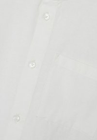 Weißes Stoffhemd mit drei sichtbaren Knöpfen und einer aufgenähten Brusttasche auf der rechten Seite.