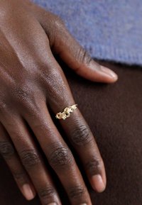 Anillo de oro con un diseño entrelazado que presenta pequeños acentos redondos, llevado en una mano contra un fondo oscuro, mostrando la textura suave de la piel.