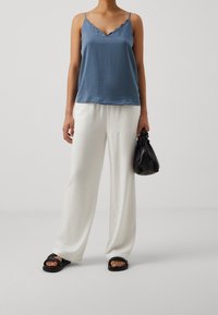 Top blu scollato con bordi ondulati, pantaloni bianchi a gamba larga, borsa nera e sandali. Tessuto morbido, vestibilità rilassata, design minimalista, colori tenui.