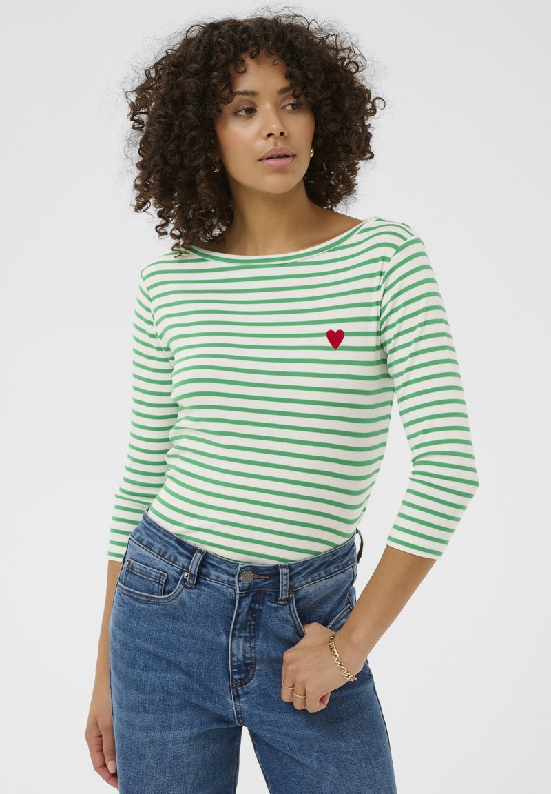 Vrouw met krullend haar, gekleed in een groen-wit gestreept shirt met lange mouwen en een rood hart-embleem, blauwe spijkerbroek en een gouden armband, kijkend naar rechts.