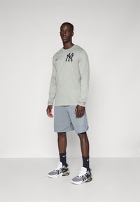 Langermet grå treningsskjorte med marineblå NY-logo, sammen med lysegrå shorts og svart-hvite joggesko. Enkelt bakgrunn.