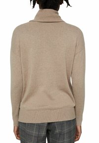 Pull à col roulé beige en maille avec une coupe décontractée, poignets et ourlet côtelés, doté d'une texture douce. Porté avec un pantalon à carreaux.