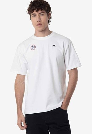 Joven con pelo rizado y oscuro que lleva una camiseta blanca de manga corta con dos pequeños logos en el pecho y pantalones negros.