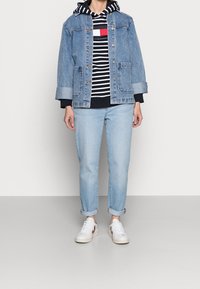Chaqueta de mezclilla con dos bolsillos frontales, una sudadera a rayas debajo, jeans de color azul claro y zapatos blancos con acentos en marrón.