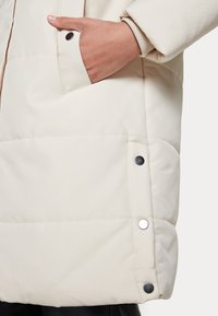 Giacca piumino beige con finitura testurizzata, dotata di tasca laterale e bottoni a pressione in argento. Design trapuntato visibile sul tessuto.