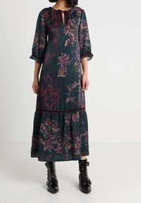 Lång, mörkgrön klänning med blommönster i rosa och lila. Har trekvartsärmar och ett tärningsliknande slut. Parat med svarta snörkängor.