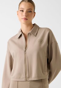 Krótka, beżowa kurtka na zamek z kołnierzem, wyposażona w przedni zamek błyskawiczny i subtelne logo. Gładki materiał, luźny krój oraz boczne kieszenie.
