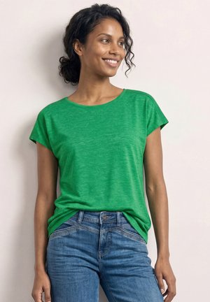 Ung kvinde med krøllet hår bundet tilbage, iført en lysegrøn kortærmet trøje og blå jeans, smilende mens hun kigger til højre.