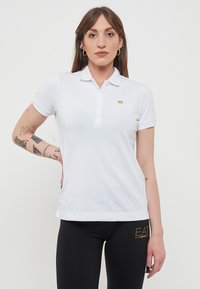 Polo shirt bianco con maniche corte, patta con tre bottoni e un piccolo logo dorato; indossato con leggings neri con un accent logo.