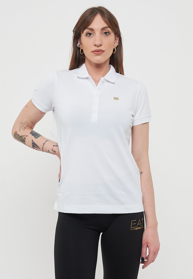 Polo shirt bianco con maniche corte, patta con tre bottoni e un piccolo logo dorato; indossato con leggings neri con un accent logo.