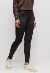 Musta legginsit joustavasta kankaasta, jossa on sileä tekstuuri, korkea vyötärö ja tiukka muotoilu valkoisten tennareiden kanssa.