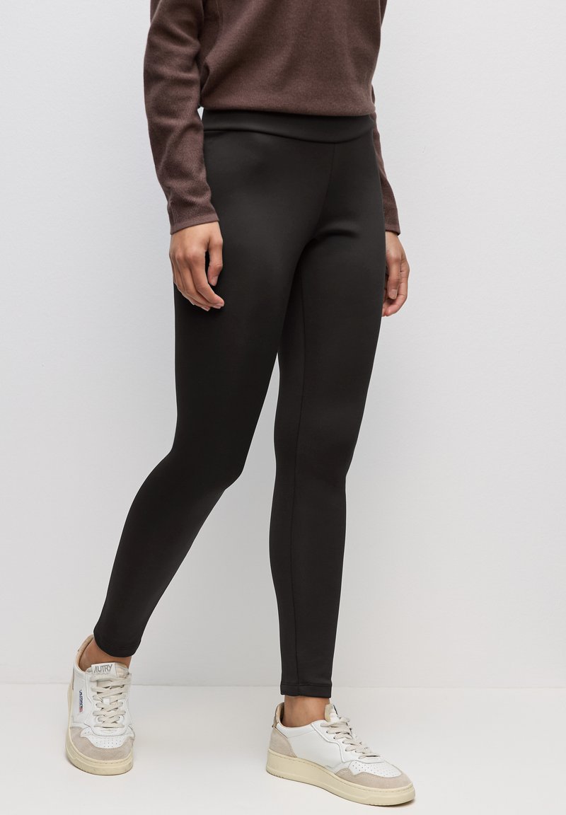 Musta legginsit joustavasta kankaasta, jossa on sileä tekstuuri, korkea vyötärö ja tiukka muotoilu valkoisten tennareiden kanssa.