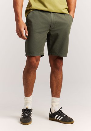 Man draagt olijfgroene korte broek, witte sokken en zwarte Adidas sneakers, staat met één hand in zijn zak.