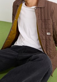 Osoba w brązowej pikowanej kurtce, nosząca białą koszulę i czarne spodnie, siedzi na zielonej podłodze.