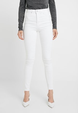 Vrouw die een witte skinny jeans met hoge taille, een grijs top met lange mouwen en witte pumps met spits toelopende neus draagt, staand tegen een effen achtergrond.