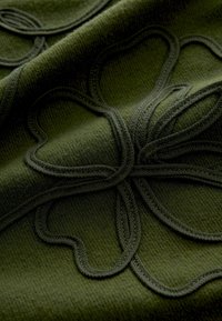 Zielona tkanina z wypukłym, plecionym wzorem kwiatowym. Teksturowana powierzchnia o różnorodnych odcieniach zieleni, podkreślająca misterny wzór.