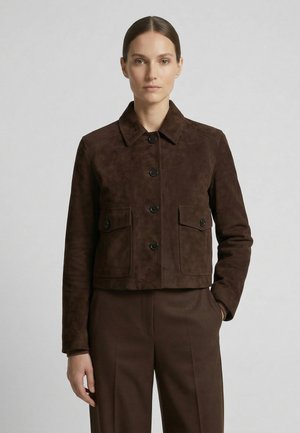 Frau trägt eine dunkelbraune Wildlederjacke mit Knopfleiste und Klappentaschen, kombiniert mit passenden braunen Hosen, vor einem schlichten grauen Hintergrund.