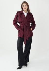 Bordowy płaszcz typu trencz z paskiem w talii, dużymi klapami i guzikami jako akcentami. Połączony z ciemnymi spodniami z wysokim stanem i butami na obcasie.