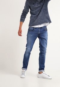 Jeans de mezclilla azul con un corte slim, que presentan un leve desgastado y diseño de cinco bolsillos, combinados con una camisa de manga larga oscura y zapatos deportivos blancos.