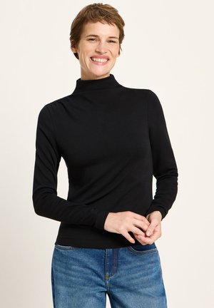 Smiling person with short hair wearing a black turtleneck and blue jeans, standing against a plain light background.