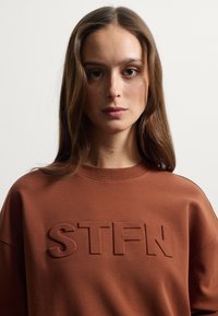 Felpa marrone con lettering in rilievo "STFN" sul davanti. Tessuto morbido, scollo rotondo e vestibilità rilassata. Design semplice senza motivi aggiuntivi.