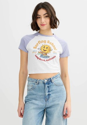 Mujer con una camiseta raglán corta blanca y lavanda con un gráfico de un sol sonriente surfeando y el texto "Escuela de Surf, Long Beach, California".