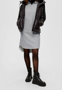 Chaqueta negra acolchada, vestido de sudadera gris con dobladillo asimétrico, medias negras y botas de combate negras de suela gruesa. Materiales suaves y texturizados.