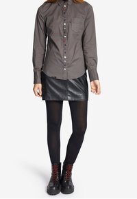 Grå skjorte med knapper og brystlomme, kombineret med en sort lædernederdel, sorte strømpebukser og sorte støvler med røde snørebånd.