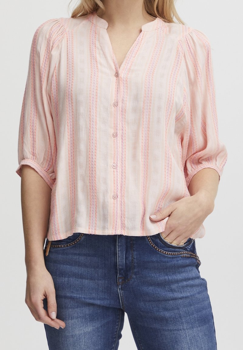 Blusa rosa chiaro con strisce verticali in tonalità di rosa più scure. Presenta un collo alla cinese, chiusura frontale a bottoni e maniche a sbuffo.