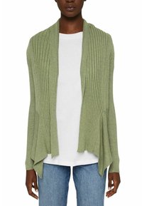 Femme portant un cardigan vert clair à côtes ouvert sur le devant, par-dessus une chemise blanche et un jean bleu, debout les bras détendus le long du corps.