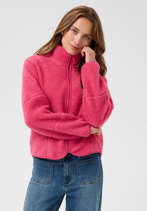 Jeune femme aux cheveux longs portant une veste polaire rose vif zippée et un jean bleu, posant avec une main près du visage.