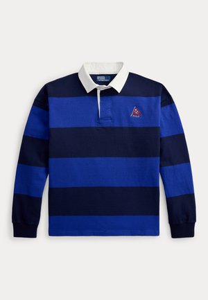 Žena a muž majú na sebe modré a tmavomodré pruhované rugby tričká; žena kombinuje rifle a hnedé mokasíny, muž biele nohavice a modré slip-ony.