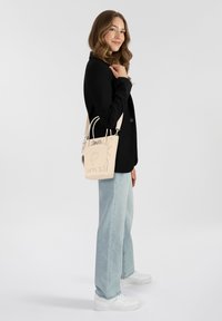 Beige Handtasche mit strukturiertem Design, ausgestattet mit zwei Tragen, einem abnehmbaren Schulterriemen und dekorativen Details wie Druckakzenten und Nieten.