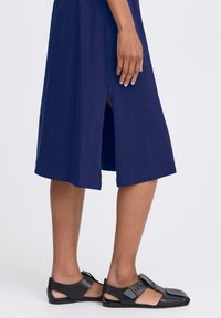Navy blue knee-length dress with a side slit, made of smooth fabric, paired with black sandals featuring an adjustable strap and open design.