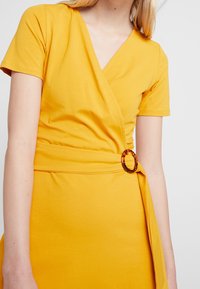 Gul omslagsklänning i mjukt tyg, med korta ärmar, V-halsringning och en framträdande ring i sköldpaddsmönster vid midjan.
