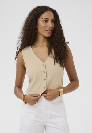 Kvinde med langt krøllet hår iført en beige ærmeløs skjorte med knapper, hvide bukser, gyldne armbånd, halskæde og øreringe, der ser til siden.