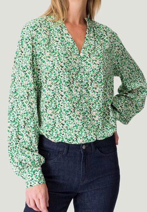 Kvinde iført en knappet, langærmet bluse med grøn, sort og beige blomsterprint, kombineret med mørkeblå jeans.