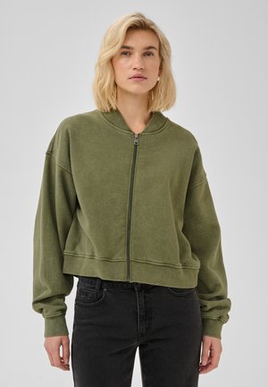 Žena s krátkymi blond vlasmi, oblečená v zelenú mikinu na zips a čierne džínsy, na jednofarebnom sivom pozadí.