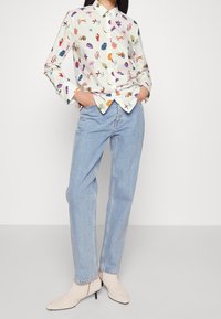 Camisa de manga larga con fondo crema y patrones abstractos coloridos, combinada con jeans de mezclilla azul claro y tacones beige con textura.