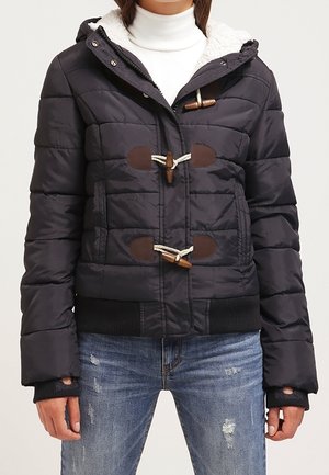 Woman wearing a black quilted jacket with toggle closures, a fur-lined hood, white turtleneck, and distressed blue jeans.