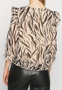 Gennemsigtig beige bluse med sorte zebrastriber og polkaprikkker, med flæseærmer og en peplum-udskæring. Lukning med knapper bagpå er synlig.