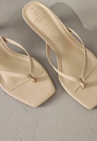 Sandales beiges avec un design à bout carré, dotées de fines straps croisées et d'une texture en cuir lisse. Marquage simple sur la semelle.