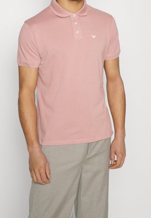 Homme portant un polo à manches courtes rose clair avec des boutons et un pantalon beige clair, debout devant un fond uni.