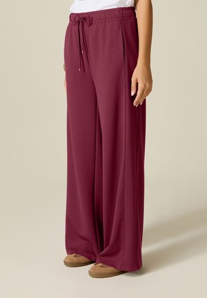 Pantalon bordeaux à jambes larges avec taille à cordon et poches latérales, porté avec des chaussures beige à enfiler sur fond uni.