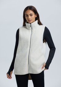 Fleece vest in off-wit met hoge kraag, volledige rits en zijzakken; gelaagd over een zwart T-shirt met lange mouwen, met een zachte textuur.